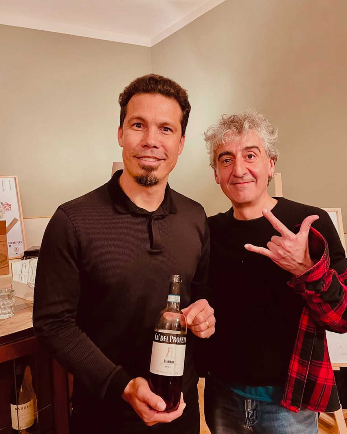 Due uomini sorridono, uno con barba tiene una bottiglia di vino rosso "Saudade Ca' del Profeti", l'altro con capelli grigi fa un gesto rock. Sfondo interno. Due uomini sorridono, uno con barba tiene una bottiglia di vino rosso "Saudade Ca' del Profeti", l'altro con capelli grigi fa un gesto rock. Sfondo interno.