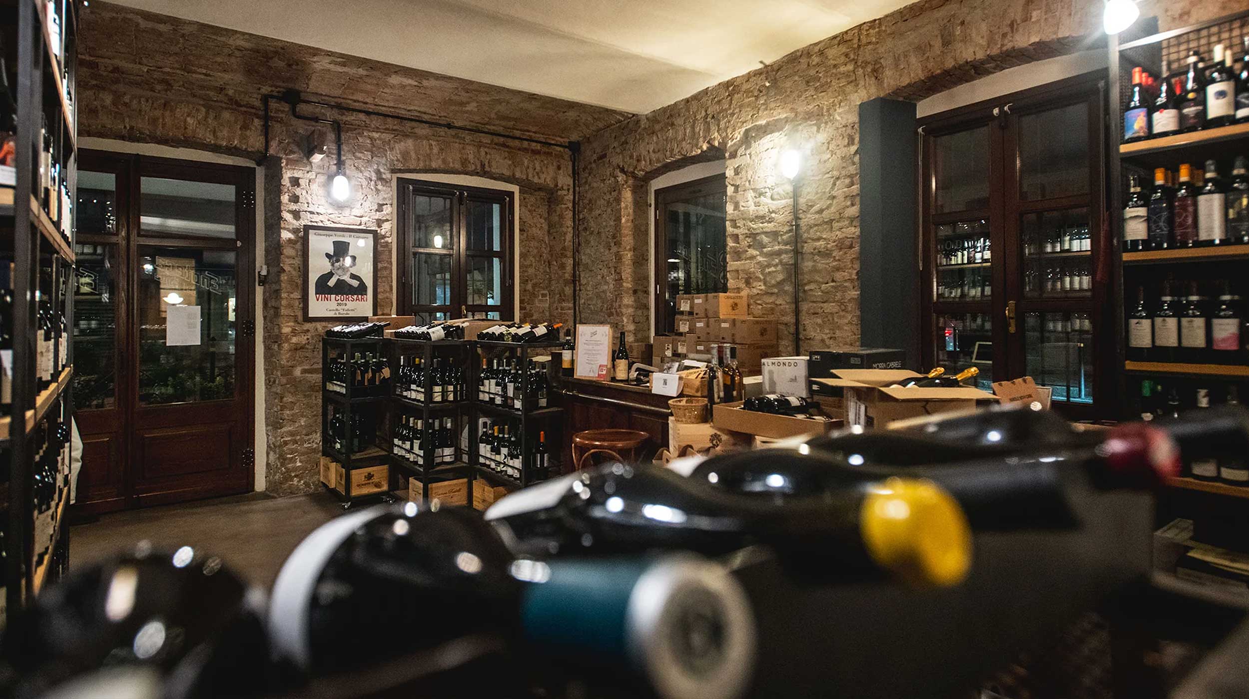 Interno di un'enoteca con pareti in mattoni a vista, scaffali e casse di vino. Bottiglie in primo piano sfocate. Poster "VINI CORSARI". Interno di un'enoteca con pareti in mattoni a vista, scaffali e casse di vino. Bottiglie in primo piano sfocate. Poster "VINI CORSARI".