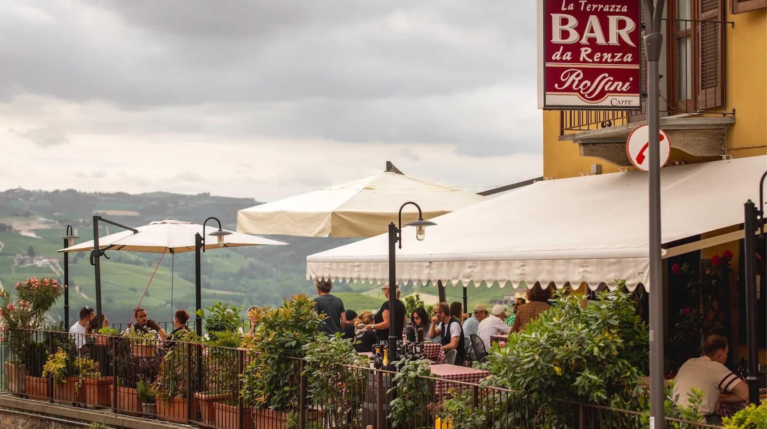 Terrazza di un bar-ristorante con persone che pranzano sotto ombrelloni, affacciata su colline verdi. Insegna "BAR da Renzo Roffini". Cielo nuvoloso. Terrazza di un bar-ristorante con persone che pranzano sotto ombrelloni, affacciata su colline verdi. Insegna "BAR da Renzo Roffini". Cielo nuvoloso.